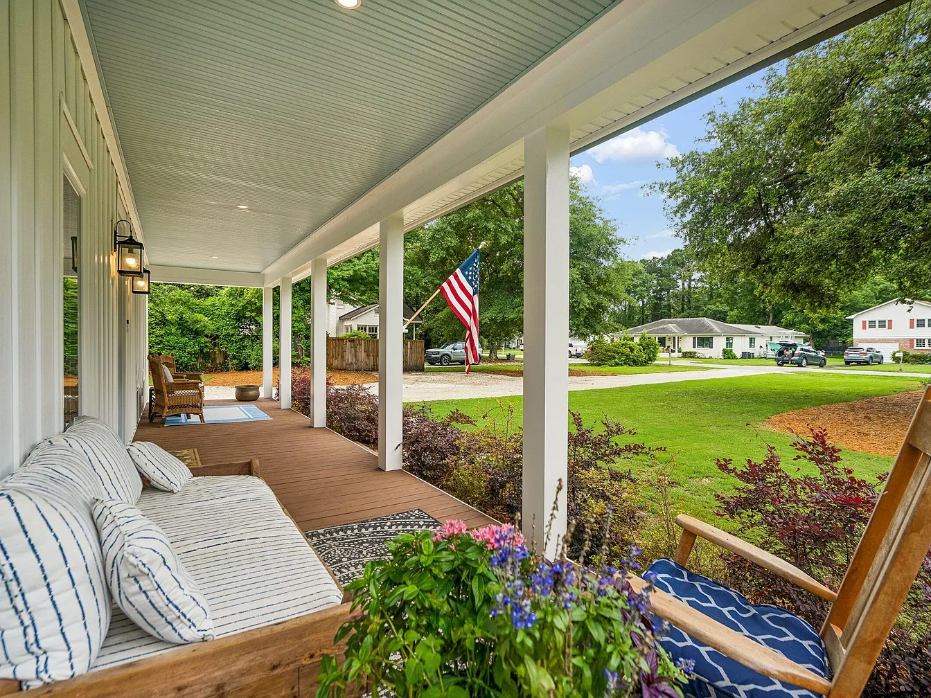 The covered front porch at 1144 Pleasant Pines, with a striped cushioned swing daybed, an American flag, painted blue ceiling, and a view of the front lawn and street beyond