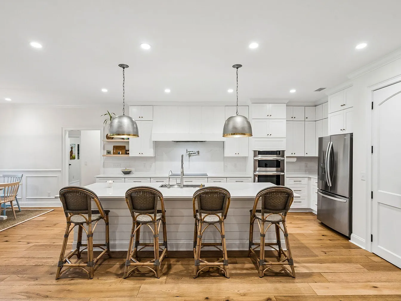 The kitchen viewed straight on across the marble island, twin polished steel pendants suspended on chains, wide-plank oak flooring leading to the dining area beyond