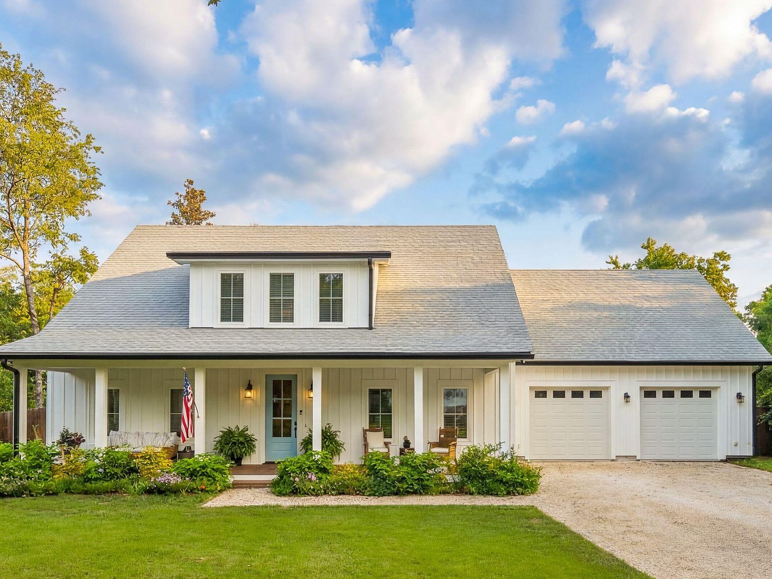 1144 Pleasant Pines photographed head-on in late-afternoon Lowcountry light, the front yard centered, twin garage doors at right, blue front door framed by potted plants, the porch flag catching the western sun, the roofline glowing against a soft southern sky