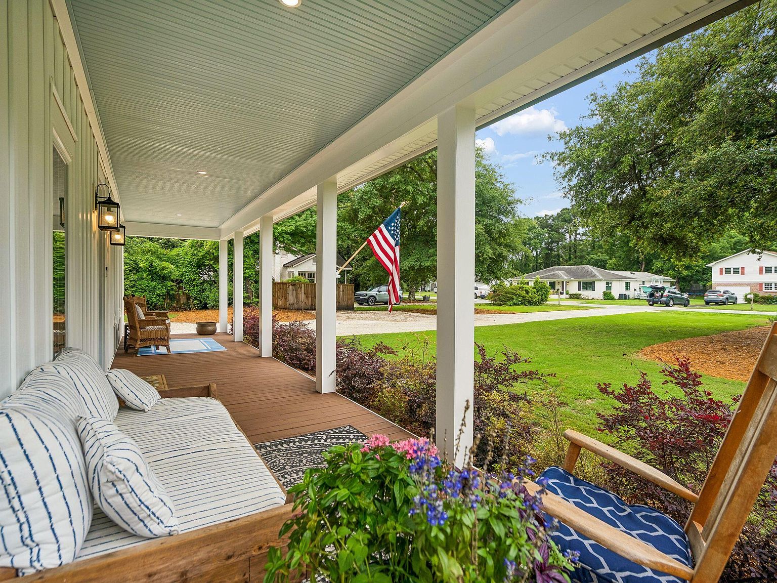 The porch at 1144 Pleasant Pines viewed from the swing daybed end: the blue painted ceiling continues across the full length, the rattan club chairs sit grouped at the far end, the American flag hangs in the middle distance, the lawn and street beyond
