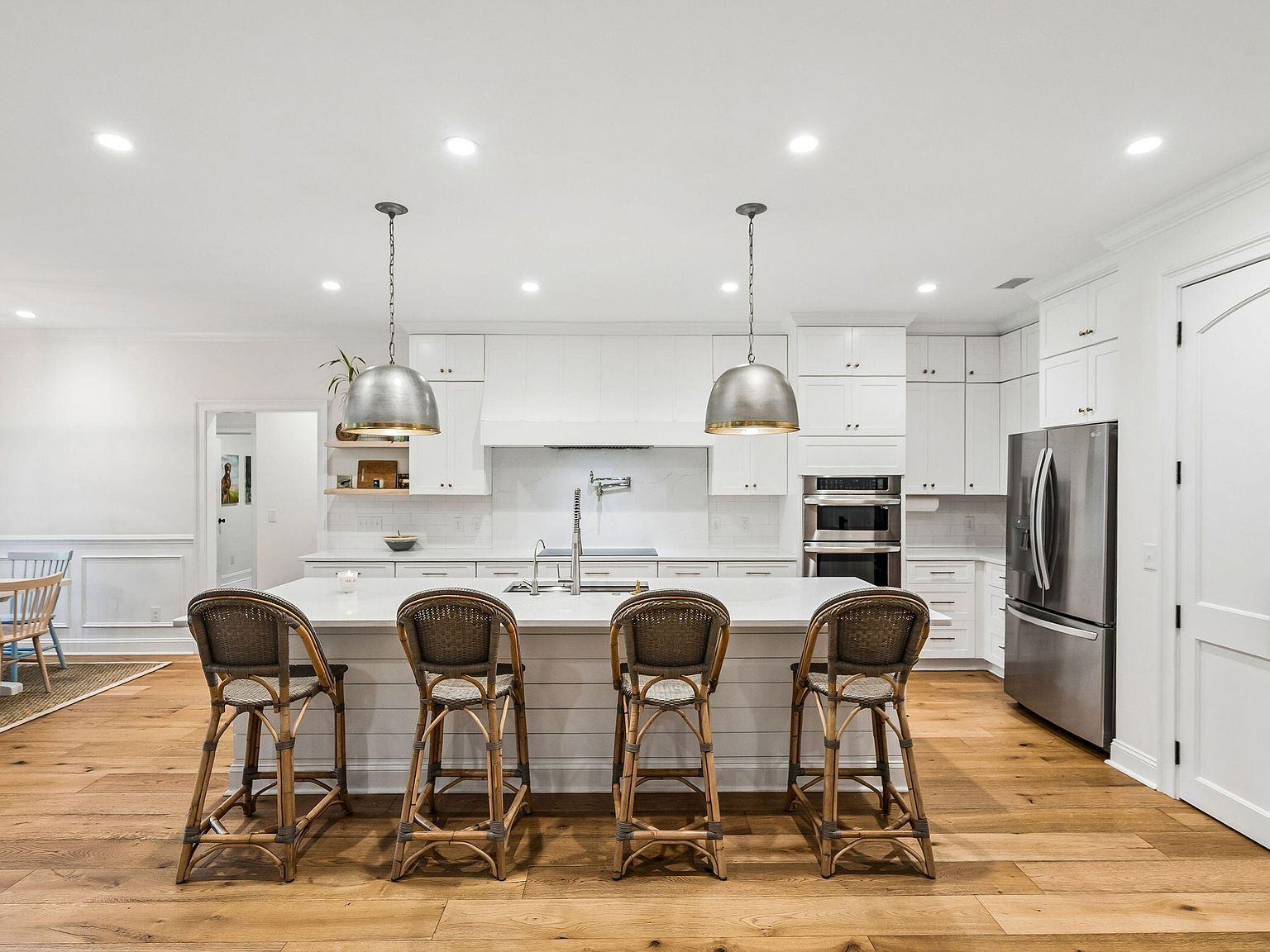 The kitchen at 1144 Pleasant Pines pulled wider, with the island and four rattan stools in the foreground, the dining room visible past the cooktop wall, a wood-bead chandelier over a long farmhouse table, and west-facing windows letting in afternoon light from the back yard