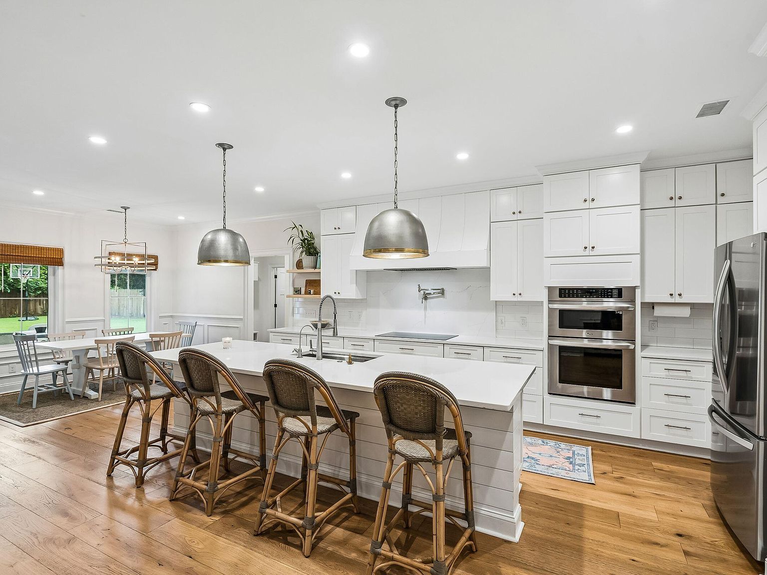 The kitchen at 1144 Pleasant Pines from the dining-room side, looking back across the island to the cooktop wall and the marble slab, four rattan stools lined up at the seating side, wide-plank oak floor continuous between rooms