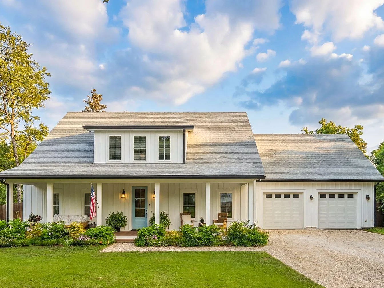 Front elevation of 1144 Pleasant Pines, a 2022 custom-built two-story Lowcountry farmhouse with white siding, a wide covered porch, twin dormers, and mature oaks in Mount Pleasant, South Carolina